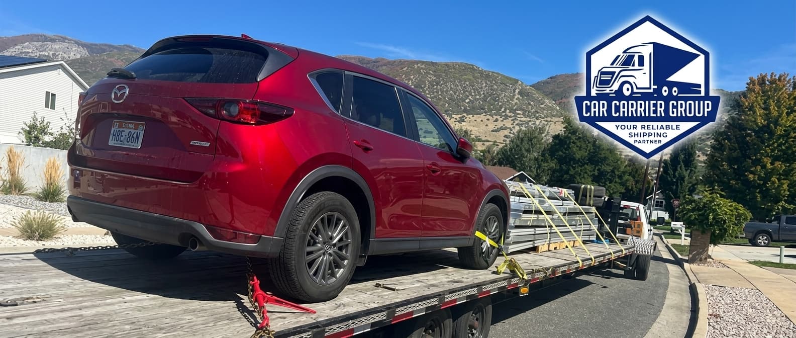 Image Description: A red SUV rests on a flatbed car trailer outdoors under a clear blue sky with mountains in the background. In the top right corner, the blue and white "CAR CARRIER GROUP" logo displays the text "CAR CARRIER GROUP" and "YOUR RELIABLE SHIPPING PARTNER